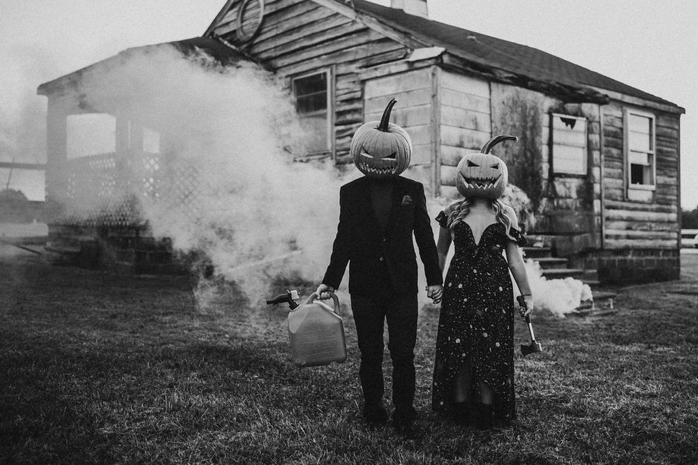 A man & woman dressed in elegant black attire have sinister grinning pumpkins on their heads. They stand in front of a dilapidated house, that appears to be on fire. The man holds a gas cannister, the woman an axe.