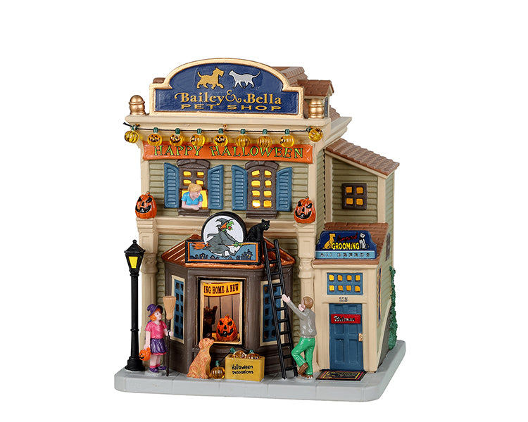 A main street shop has a large blue sign that reads Bailey & Bella Pet Shop and below that an orange sign that reads Happy Halloween. There is a man climbing a ladder to fetch a black cat on the roof and a woman assisting by peaking hear head out the second story window. A small girl wearing a witch costume is out front with a dog. 