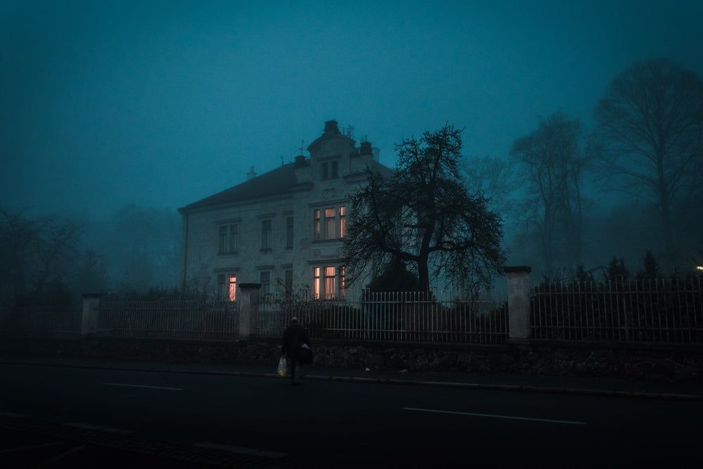 A gothic mansion is surrounded by a cast iron fence, spooky woods and a blueish black evening sky. A few windows have an eerie yellow glow to them as a man appears to be returning from work to head inside.