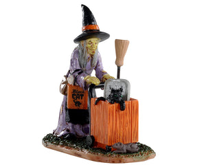 A witch wearing a black and orange hat and purple dress pushes an orange cat with a black cat inside. She also has a broom and various shopping bags.