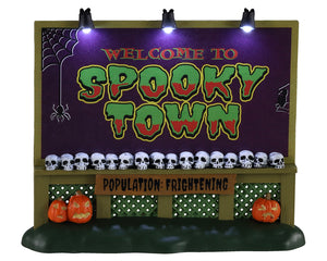 A Spooky lighted sign with the words Welcome To Spooky Town and population frightening is illuminated by overhead lights. 3 jack-o'-lanterns sit at the base of the sign while a long row of skulls sits right beneath the words Spooky Town on the center bottom of the sign.