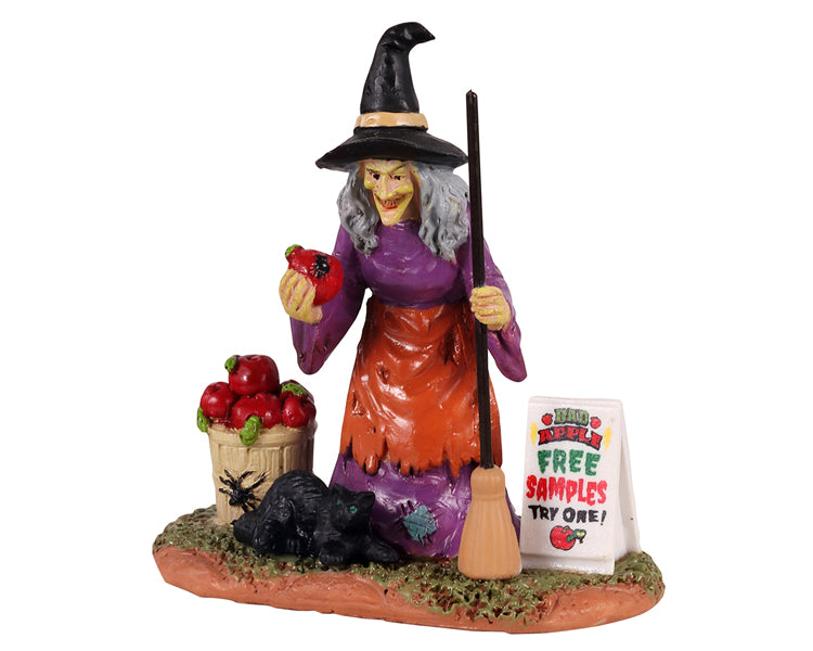 An evil witch holds a poisonous apple in her right hand and a broom in her left while her black cat, basket of rotten apples and a sign reading FREE SAMPLES sit at her feet.