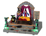 Two Witches perform a levitation on a woman in a yellow dress. They're standing on a stone stage with steps covered in blood, bones, rats and gargoyles.