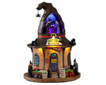 A witch hat shaped building called Helga's Hats & More has multiple witches working on creating hats. The building has multiple yellow and blue colored lights.