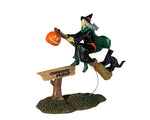 A witch dressed in black with a green cape rides her broom over a small sign that reads curbside pickup. She has a pumpkin basket on the front of her broom and her black cat sidekick on the back.