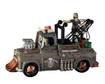 A rusty mad max style tow truck is driven by a green ghoul with another monster riding in the back of the truck. The side of the truck says, "24/7 Towing & Salvage"