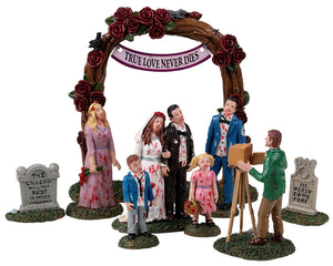 A zombie wedding party poses for pictures while their outfits are covered in blood. They're standing between tombstones and there's 5 adults (including the photographer) and 2 children.