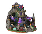 A large amusement park ride sits at the entrance to a cave. At the top of the ride is a sign that reads, "The Count's Bat Bonanza" and just below that is Dracula overlooking at the riders. The riders are sitting inside purple bat seats that spin around the center tower. There's purple and green lighting and coffins. 