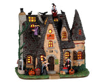 A wicked witch house is crawling with witches, black cats and crows. It has a funky witch roof that is covered in black and grey shingles and there's a yellow light coming from the building.
