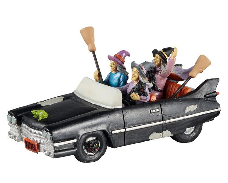This table piece is a black Halloween-themed convertible with black cat rims. Inside the car are three witches enjoying their joy ride around town while they hold onto their trusty black cats and brooms. The witches are dressed in their standard witches' hats and capes. A green toad sits on the hood of the car, joining along for the ride.