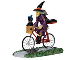 A witch dressed in purple with a black and orange hat and cape rides a red bicycle, while her black dog rides in the basket up front. 