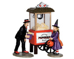 A well dressed mustached man with a top hat offers popcorn from his popcorn machine to a young trick-or-treater girl dressed as a witch.