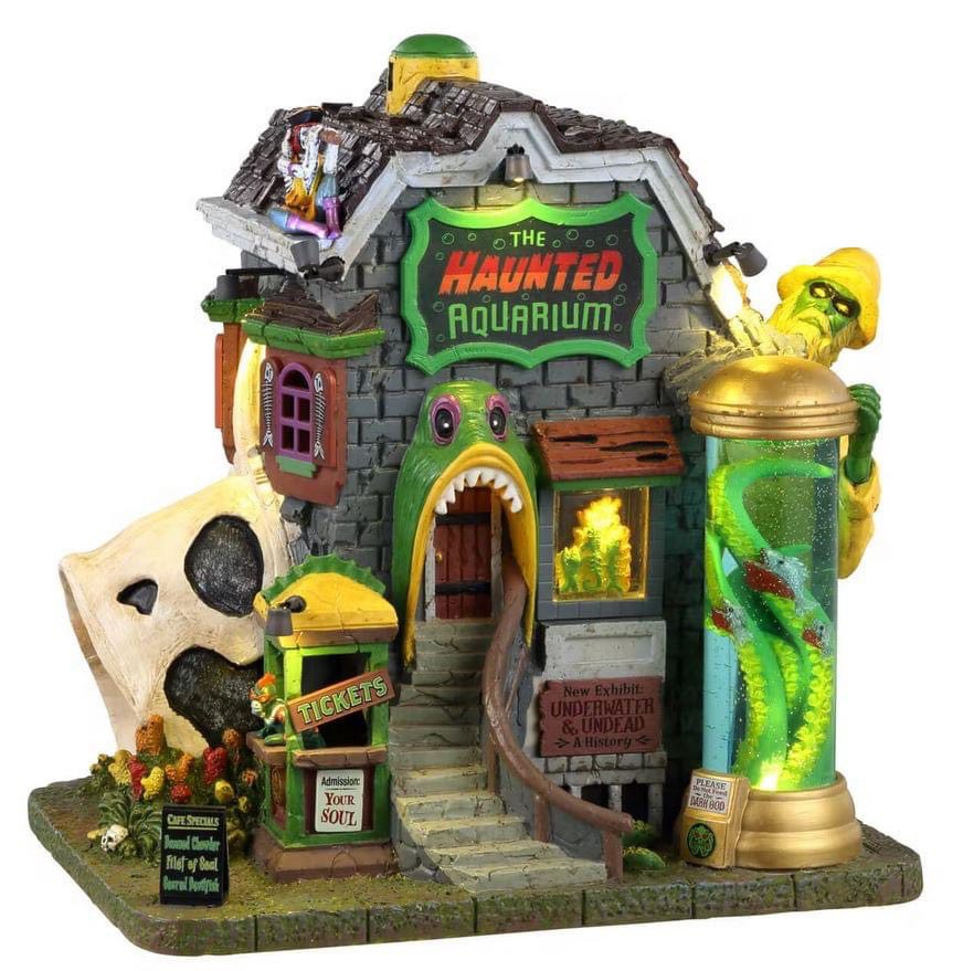 Lemax Spooky Town Haunted Aquarium #35001 is a building covered in green fish and a monster boat captain. A green and yellow ticket booth is out front on the left, while a large cylinder is home to eel sea monsters on the right side of the building.