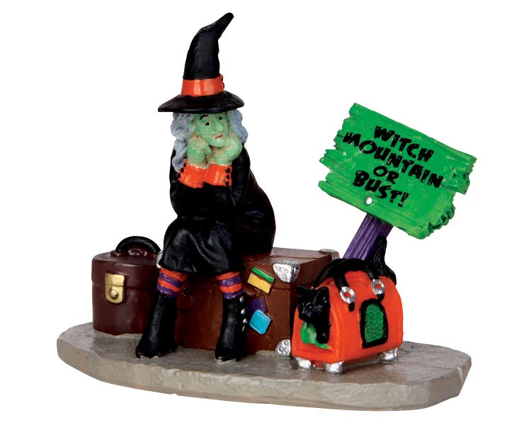 A witch is stranded on the sidewalk. She sits on a trunk surrounded by other pieces of luggage and a green sign that reads "Witch Mountain or Bust!". A black cat peaks out of one of her bags.
