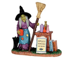 A witch stands over a table covered in potion bottles while she holds a broom and her black cat stands next to her.