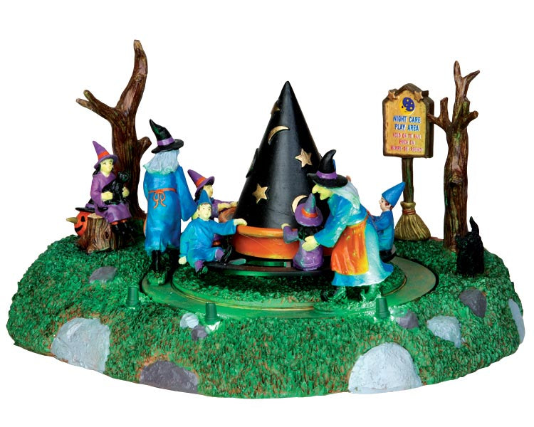 Two adult witches play with 6 children witches who spin around a witch hat shaped ride, all are dressed in blue and purple. Two trees and a sign reading night care play area sit behind the group. Multiple black cats also hang out with their  witches.