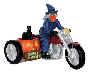 A witch dress in blue with an orange face mask bandana drives a motorcycle while her black cat rides in a pumpkin sidecar. 