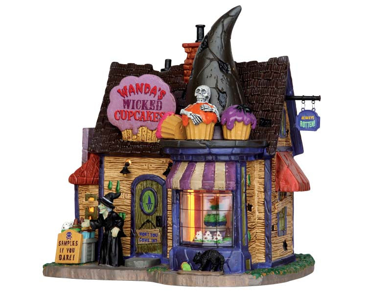 A small wood house with a shingled roof has a purple and pink sign on top of it that reads Wanda's Wicked Cupcakes. A witch stands out front while both witchy and cupcake accents covered the building.