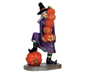 A friendly witch dressed in purple rests one leg on top of a large pumpkin while she holds a stack of 5 smaller pumpkins in her hands.