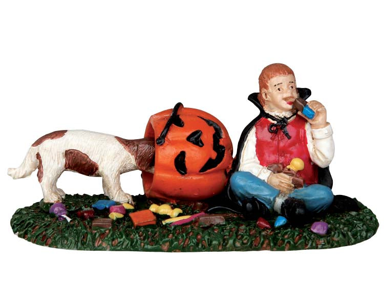 A candy crazed trick-or-treater boy dressed as a vampire sits on the ground and indulges in a chocolate bar while his naughty puppy sneaks candy from a toppled over pumpkin candy bucket. 