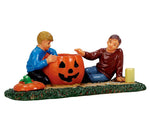 A dad lays next to his son and teaches him how to properly carve his first jack-o'-lantern while the son eagerly scrapes away any remain pumpkin insides. 