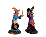 Two witches, one dressed in blue and the other in purple decide on hats to wear. The witch in purple has a black dog sidekick that is wearing an orange witch hat. 