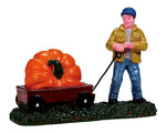 A man in a yellow jacket and blue hat wheels a massive pumpkin in a red wagon.