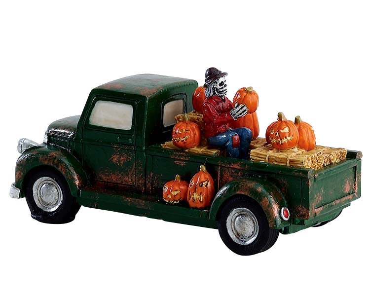 A skeleton scarecrow man sits on haybales in the back of his green truck. He holds one jack-o'-lantern and is surrounded by a bunch more. 