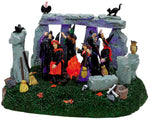 Joyful Witches dance around a fire in the center of Stonehenge, while various forest birds and a black cat look on. 