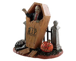 A zombie comes back from the dead by breaking out of their coffin which is leaning up against a small metal fence and a jack o' lantern. 
