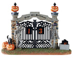 A gothic stone and iron fence topped by pumpkins is guarded by two skeletons wearing black and purple top hats. A sign on top of the fence reads Days Til Halloween with the number of days listed below.