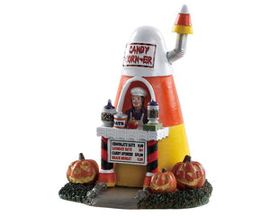 A man that looks like a vampire sells various candy out of a small structure that has the shape and color of a piece of candy corn. Three jack-o'-lanterns sit in front of the sturucture.