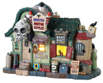 A Small building with a sign that reads "Monster Hunting Supplies" is covered in various supplies like garlic, explosives, weapons and more. The roof is green and there is a large skull over the door while a yellow glow emits from the inside.