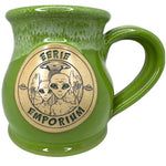 Two aliens appear on a bright green drip glaze coffee mug in front of their UFO. EERIE EMPORIUM is written in green halloweeny font.