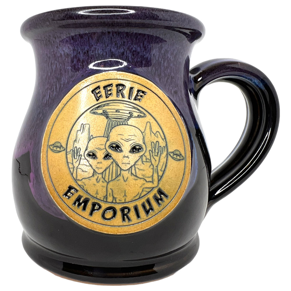 Two aliens appear on a blue, blue and black drip glaze coffee mug in front of their UFO. EERIE EMPORIUM is written in purple halloweeny font.
